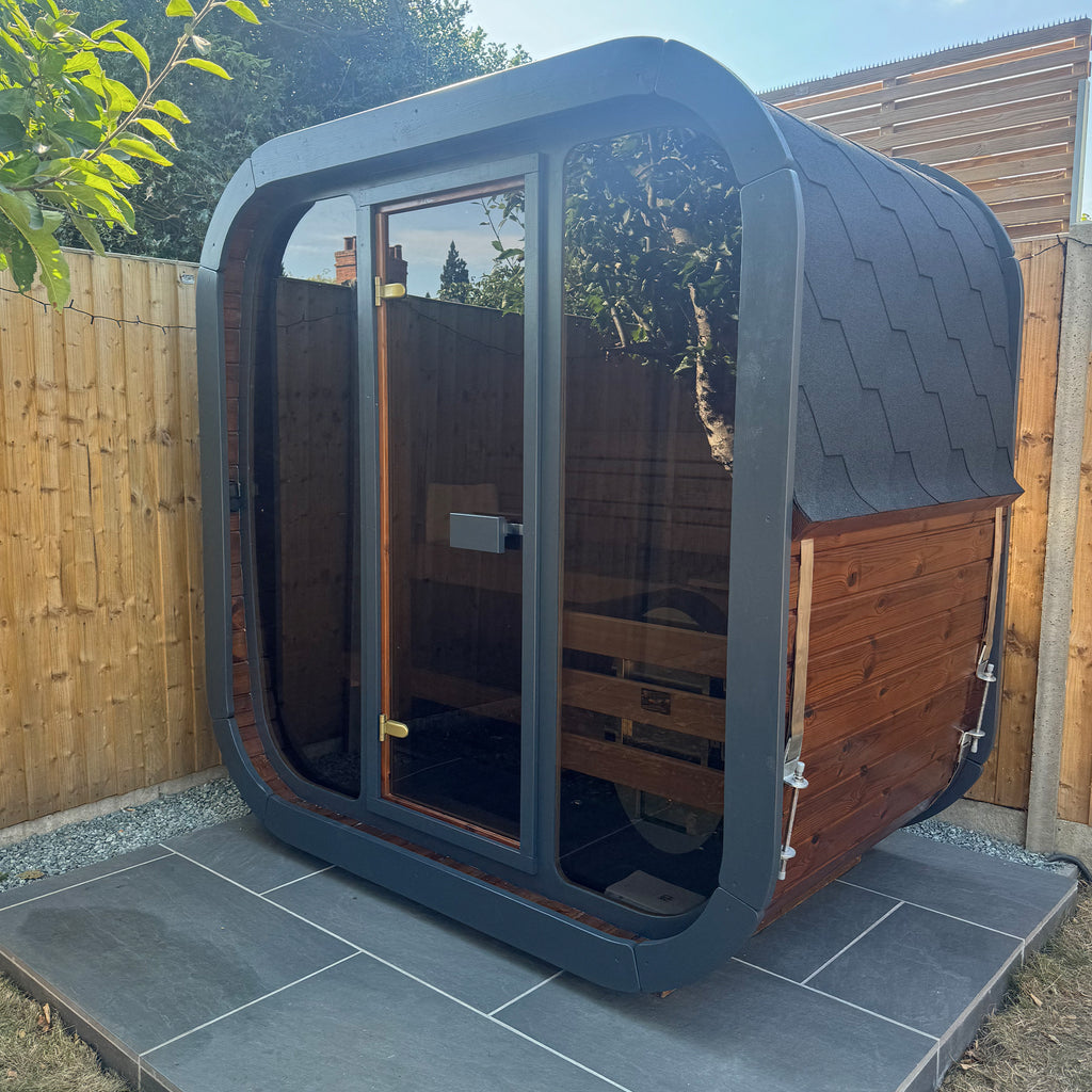 Modern outdoor Sauna cabin KC125 with glass walls on a tiled platform, surrounded by wooden fence and trees.