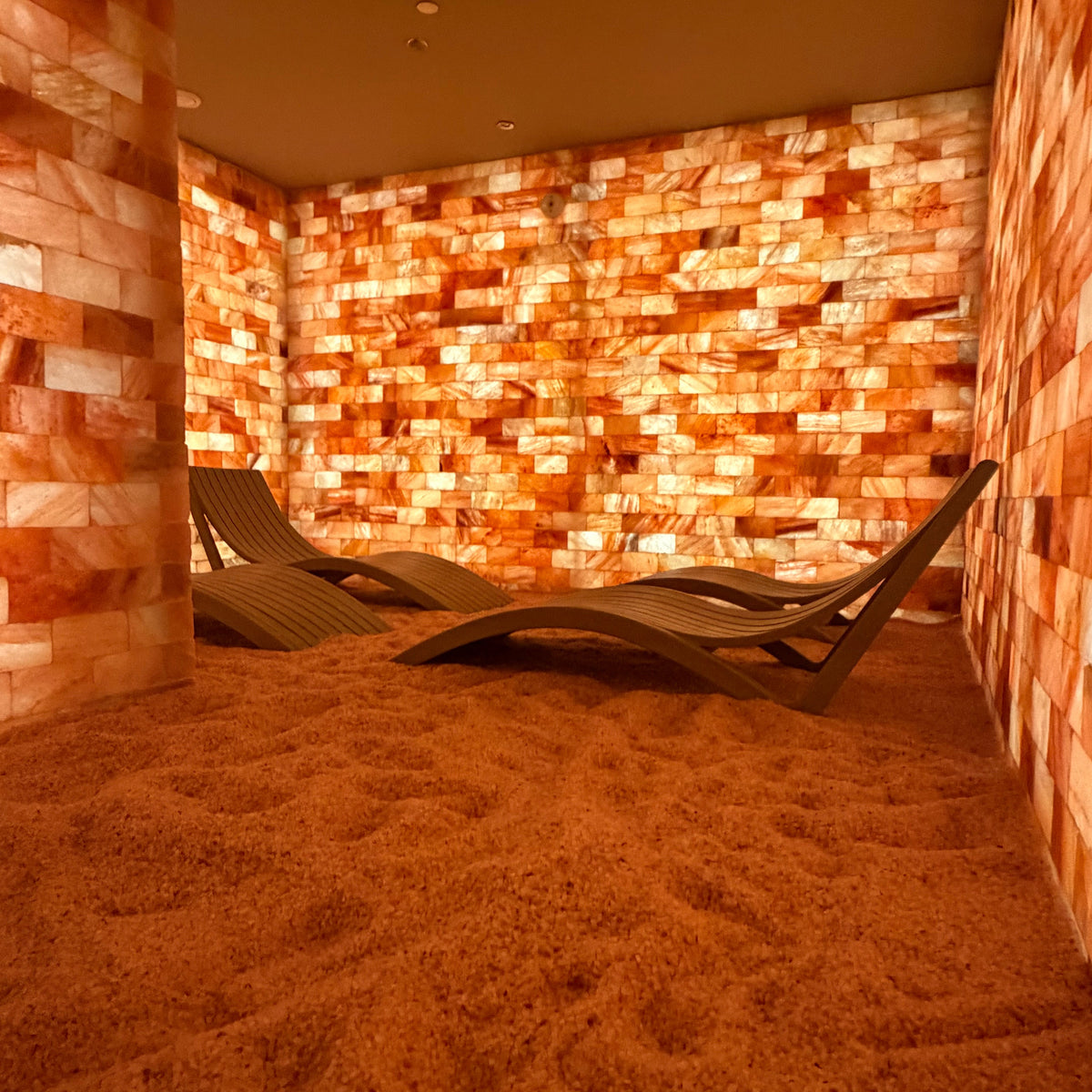 Design reference: illuminated pink and amber brick walls, a granular salt floor and curved slatted loungers in a salt room.